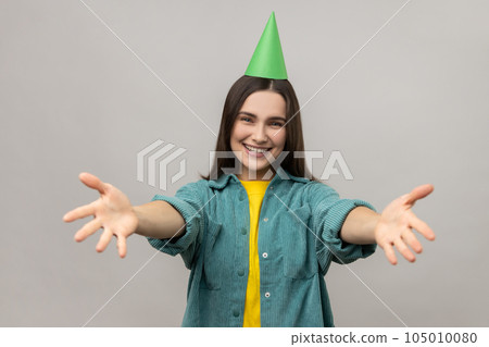 Woman with party cone keeping hands wide open to embrace, giving warm welcome, greeting and sharing love, wearing casual style jacket. Indoor studio shot isolated on gray background. 105010080