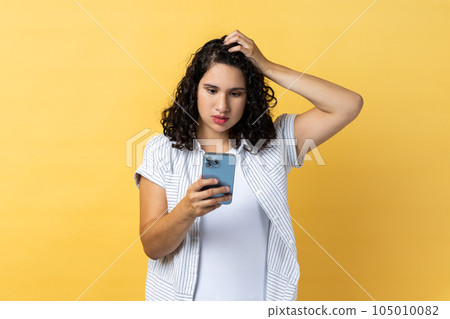 Portrait of pensive beautiful young adult woman with dark wavy hair using cell phone thinking what to answer, keeps hand on head. Indoor studio shot isolated on yellow background. 105010082
