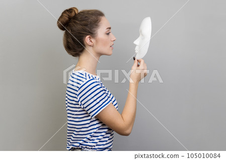 Side view portrait of beautiful dark haired woman with bun hairstyle wearing striped T-shirt standing holding and looking at white mask. Indoor studio shot isolated on gray background. 105010084
