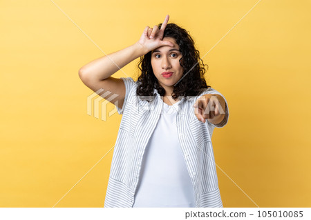 Portrait of grumpy disrespectful woman with dark wavy hair showing loser gesture and pointing finger at camera, blaming and mocking. Indoor studio shot isolated on yellow background. Portrait of grumpy disrespectful woman with dark wavy hair showing loser gesture and pointing finger at camera, blaming and mocking. Indoor studio shot isolated on yellow background. 105010085