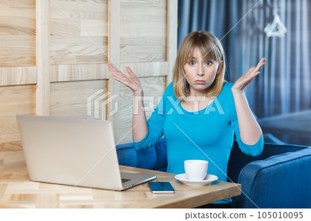 Confused uncertain young woman with blonde hair in blue shirt sitting at table and working on laptop, shrugging shoulders, looking at camera with puzzled face. Indoor shot, cafe background. 105010095