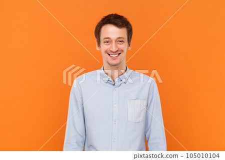 Portrait of overjoyed positive happy man standing looking at camera with toothy smile, expressing happiness, wearing light blue shirt. Indoor studio shot isolated on orange background. Portrait of overjoyed positive happy man standing looking at camera with toothy smile, expressing happiness, wearing light blue shirt. Indoor studio shot isolated on orange background. 105010104