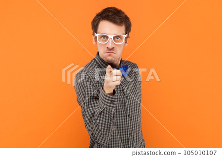 Self assured serious man nerd points index finger at camera chooses you looks directly at camera, wearing shirt with blue bow tie and white glasses. Indoor studio shot isolated on orange background. 105010107