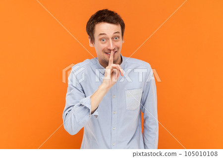 I won't tell ever. Portrait of funny positive optimistic man looking at camera and showing shh gesture, wearing light blue shirt. Indoor studio shot isolated on orange background. 105010108