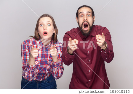 Portrait of shocked amazed woman and man standing together and pointing at camera, choosing you, looking at camera with big eyes. Indoor studio shot isolated on gray background. Portrait of shocked amazed woman and man standing together and pointing at camera, choosing you, looking at camera with big eyes. Indoor studio shot isolated on gray background. 105010115