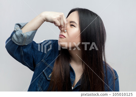 Portrait of disgusted brunette woman in blue denim jacket standing plugs nose as smells something stink and unpleasant, feels aversion. Indoor studio shot isolated on gray background. Portrait of disgusted brunette woman in blue denim jacket standing plugs nose as smells something stink and unpleasant, feels aversion. Indoor studio shot isolated on gray background. 105010125