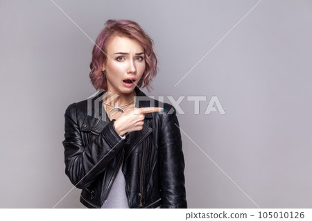 Portrait of shocked surprised woman with short hairstyle standing pointing away at copy space for advertisement, wearing black leather jacket. Indoor studio shot isolated on grey background. Portrait of shocked surprised woman with short hairstyle standing pointing away at copy space for advertisement, wearing black leather jacket. Indoor studio shot isolated on grey background. 105010126