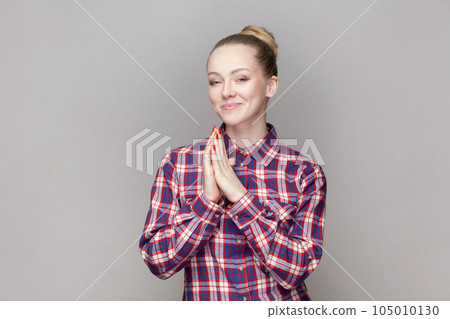 Portrait of sly attractive woman with bun hairstyle keeps palms together, planning devil plan or prank, wearing checkered shirt. Indoor studio shot isolated on gray background. Portrait of sly attractive woman with bun hairstyle keeps palms together, planning devil plan or prank, wearing checkered shirt. Indoor studio shot isolated on gray background. 105010130