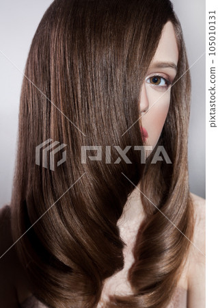 Portrait of attractive brunette woman fashion model with silk straight shiny hair and makeup, covering half of face with hair, looking at camera. Indoor studio shot isolated on gray background. Portrait of attractive brunette woman fashion model with silk straight shiny hair and makeup, covering half of face with hair, looking at camera. Indoor studio shot isolated on gray background. 105010131