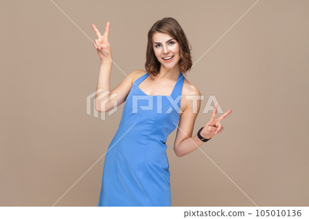 Portrait of happy positive optimistic woman with wavy hair makes peace gesture keeps lips folded shows v sign, wearing blue dress. Indoor studio shot isolated on light brown background. 105010136