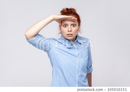 Portrait of scared ginger woman wearing blue shirt keeping palm over head and looking attentively far away, peering at long distance. Indoor studio shot isolated on gray background. Portrait of scared ginger woman wearing blue shirt keeping palm over head and looking attentively far away, peering at long distance. Indoor studio shot isolated on gray background. 105010151