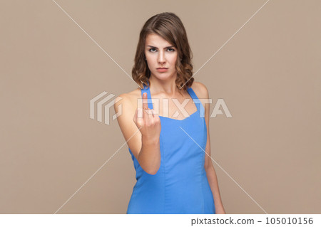 Portrait of displeased woman with wavy hair showing fuck sign, showing her negative feeling, looking at camera, wearing blue dress. Indoor studio shot isolated on light brown background. 105010156