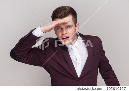 Man peering into distance, looking far away with attentive view, searching on horizon and curious to discover, wearing violet suit and white shirt. Indoor studio shot isolated on grey background. 105010157