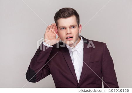 Young man holding hand near ear trying to hear secrets and find out gossips, listen to private whispers, , wearing violet suit and white shirt. Indoor studio shot isolated on grey background. 105010166