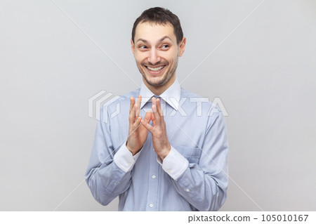 Portrait of cunning sly young adult man standing looking away with toothy smile, having devil plans, planning prank, wearing light blue shirt. Indoor studio shot isolated on gray background. 105010167