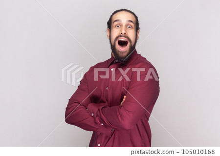 Portrait of amazed surprised man with dark hair and beard in red shirt standing with crossed arms, looking at camera with big eyes. Indoor studio shot isolated on gray background. 105010168