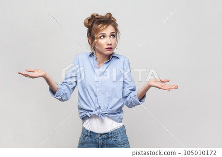 Who knows. Clueless hesitant beautiful blonde woman spreads palms sideways shrugs shoulders has indecisive expression, looking away, wearing blue shirt. Indoor studio shot isolated on gray background. 105010172