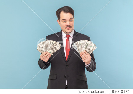 Portrait of rich man with mustache standing counting money., holding big fans of dollar banknotes, wearing black suit with red tie. Indoor studio shot isolated on light blue background. 105010175