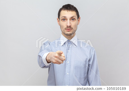 Portrait of bossy strict handsome man pointing at you with index finger, choosing or warning you, looking at camera, wearing light blue shirt. Indoor studio shot isolated on gray background. 105010189