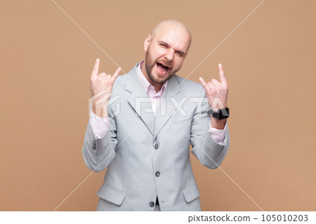 Portrait of bald bearded man rocker enjoys positive vibes, listens rock n roll, cool music in club, shows horn gesture, wearing gray jacket. Indoor studio shot isolated on brown background. 105010203