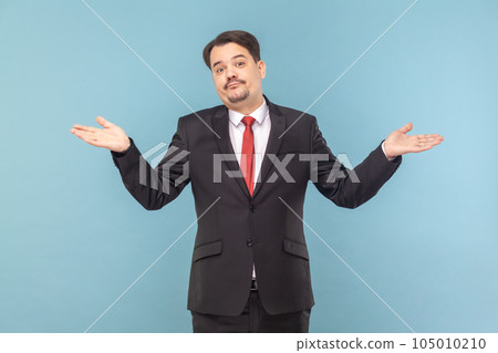 Portrait of confused uncertain man with mustache with spread hands, looking at camera, shrugging shoulders, wearing black suit with red tie. Indoor studio shot isolated on light blue background. 105010210