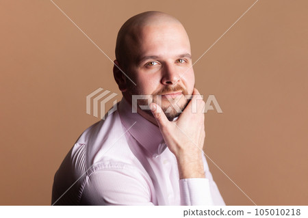 Side view portrait of cute positive smiling bald bearded man sitting holding chin, looking at camera with positive expression, wearing light pink shirt. Indoor studio shot isolated on brown background 105010218