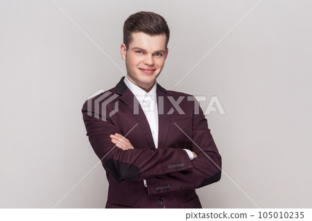 Portrait of confident clever handsome young man standing with crossed arms, looking at camera with happy expression, wearing violet suit and white shirt. Indoor studio shot isolated on grey background 105010235