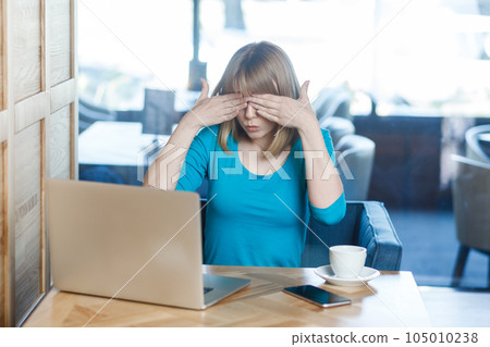 Portrait of attractive young woman with blonde hair in blue shirt working on laptop, covering eyes with palms, doesn't want to see something shameful. Indoor shot in cafe with big window on background 105010238