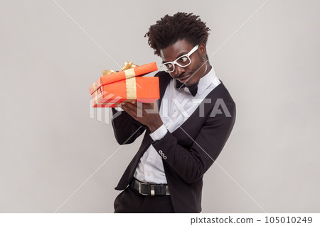 Portrait of curious handsome young adult man in glasses standing with red gift box, looking inside, wearing white shirt and tuxedo. Indoor studio shot isolated on gray background. Portrait of curious handsome young adult man in glasses standing with red gift box, looking inside, wearing white shirt and tuxedo. Indoor studio shot isolated on gray background. 105010249