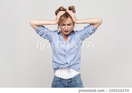 Portrait of blonde woman suffering from migraine, touching head, needs painkillers, cannot hold pain, feels discomfort, wearing blue shirt. Indoor studio shot isolated on gray background. 105010254