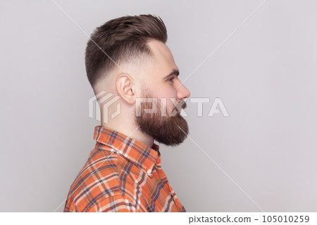 Side view portrait of serious strict bossy bearded man standing looking ahead, expressing confidence, wearing orange checkered shirt. Indoor studio shot isolated on gray background. 105010259