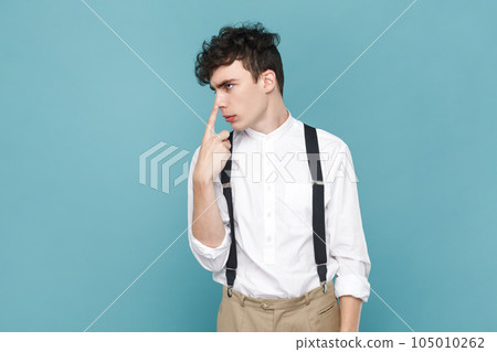 Portrait of man wearing white shirt and suspender, standing, touching his nose and showing lie gesture, suspecting falsehood, dishonest talk. Indoor studio shot isolated on blue background. 105010262