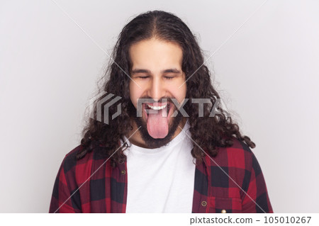 Comic bearded man in checkered red shirt sticks out tongue, makes happy grimace, expresses positive emotions, foolishes with friends. Indoor studio shot isolated on gray background. 105010267