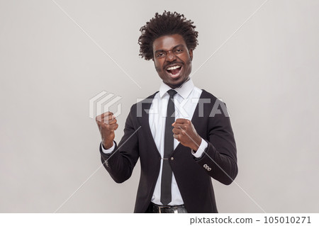 Portrait of overjoyed man with petit goatee clenches fists with joy, feels very happy and lucky, exclaims loudly, wearing white shirt and tuxedo. Indoor studio shot isolated on gray background. Portrait of overjoyed man with petit goatee clenches fists with joy, feels very happy and lucky, exclaims loudly, wearing white shirt and tuxedo. Indoor studio shot isolated on gray background. 105010271