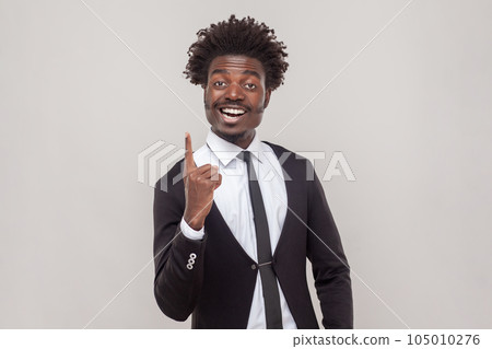 Clever man with Afro hairstyle get new idea, raises fore finger, ready to start work on project, confident in success, wearing white shirt and tuxedo. Indoor studio shot isolated on gray background. 105010276