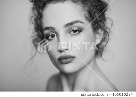Black and white beauty portrait of young confident woman fashion model with collected dark curly hair and makeup, looking at camera with calm face. Indoor studio shot isolated on gray background. 105010284