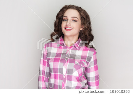 Portrait of woman with curly hair being very glad smiling with broad smile showing her perfect teeth, having fun, wearing pink checkered shirt. Indoor studio shot isolated on gray background. Portrait of woman with curly hair being very glad smiling with broad smile showing her perfect teeth, having fun, wearing pink checkered shirt. Indoor studio shot isolated on gray background. 105010293