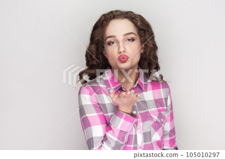 Romantic attractive woman with curly hair stretches hand near face, blows air kiss, expresses love and affection, wearing pink checkered shirt. Indoor studio shot isolated on gray background. 105010297