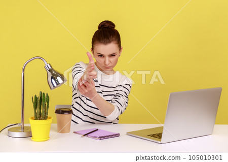Angry woman office worker sitting at workplace with laptop threatening and pointing finger gun to camera, shooting with aggressive expression. Indoor studio studio shot isolated on yellow background. Angry woman office worker sitting at workplace with laptop threatening and pointing finger gun to camera, shooting with aggressive expression. Indoor studio studio shot isolated on yellow background. 105010301