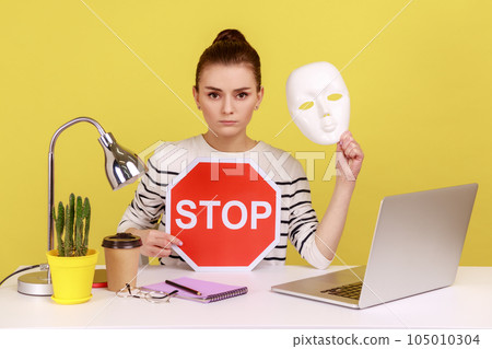 Portrait of serious dark haired woman holding white mask with unknown face and red traffic sign while sitting on workplace with laptop. Indoor studio studio shot isolated on yellow background. 105010304