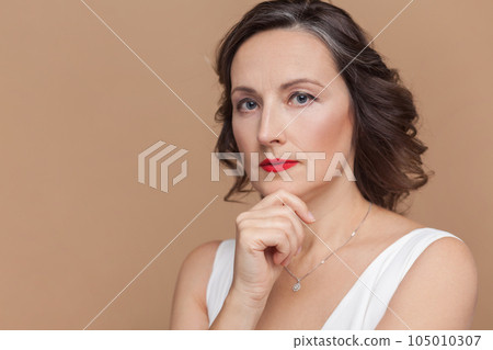 Closeup of serious pensive attractive beautiful middle aged woman with wavy hair and makeup, looking at camera, holding chin, wearing white dress. Indoor studio shot isolated on light brown background Closeup of serious pensive attractive beautiful middle aged woman with wavy hair and makeup, looking at camera, holding chin, wearing white dress. Indoor studio shot isolated on light brown background 105010307