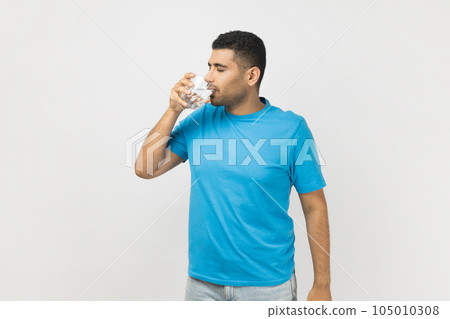 Portrait of thirsty handsome unshaven man in blue T- shirt standing with glass in hands, drinking fresh clean cold water during hot weather, enjoying. Indoor studio shot isolated on gray background. Portrait of thirsty handsome unshaven man in blue T- shirt standing with glass in hands, drinking fresh clean cold water during hot weather, enjoying. Indoor studio shot isolated on gray background. 105010308