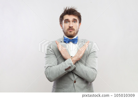 Portrait of upset attractive bearded man standing showing x sign, no way gesture, looking at camera, wearing grey suit and blue bow tie. Indoor studio shot isolated on gray background. Portrait of upset attractive bearded man standing showing x sign, no way gesture, looking at camera, wearing grey suit and blue bow tie. Indoor studio shot isolated on gray background. 105010309