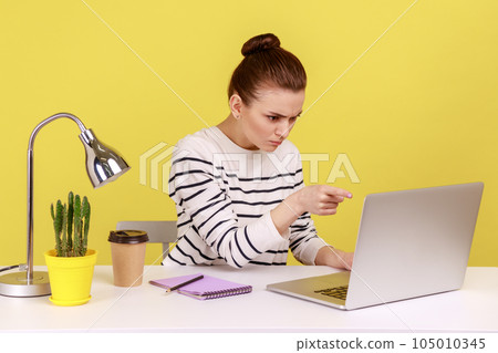 Angry woman office worker in striped shirt pointing to laptop screen, scolding and accusing talking on video call, having online conference. Indoor studio studio shot isolated on yellow background. 105010345