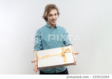 Portrait of satisfied friendly handsome young man standing with gift in hands, giving present to her girl friend on birthday, wearing blue shirt. Indoor studio shot isolated on gray background. 105010364