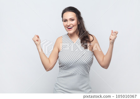 Emotional brunette woman raises clenched fists, exclaims with excitement, rejoices sweet success, feels taste of victory, wearing striped dress. Indoor studio shot isolated on gray background. Emotional brunette woman raises clenched fists, exclaims with excitement, rejoices sweet success, feels taste of victory, wearing striped dress. Indoor studio shot isolated on gray background. 105010367