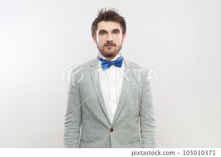 Portrait of serious attentive concentrated bearded man standing looking at camera with strict bossy expression, wearing grey suit and blue bow tie. Indoor studio shot isolated on gray background. 105010371