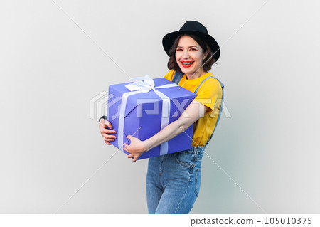 Portrait of extremely happy optimistic hipster woman in blue denim overalls, yellow T-shirt and black hat, embracing big wrapped box with present. Indoor studio shot isolated on gray background. 105010375