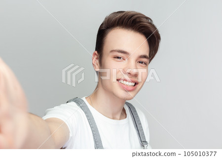 Portrait of optimistic young brunette man standing keeps arms outstretched has optimistic cheerful expression, wearing denim overalls. Indoor studio shot isolated on gray background. 105010377