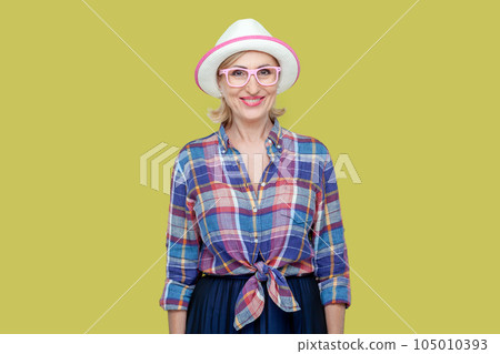 Portrait of smiling joyful senior woman wearing checkered shirt, hat and eyeglasses, looking at camera with toothy smile, expressing positive emotions. Indoor studio shot isolated on yellow background 105010393